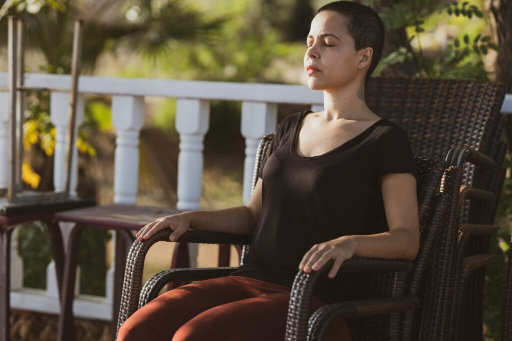 A woman in meditation with eyes closed sitting on a chair outdoors in the sunlight.