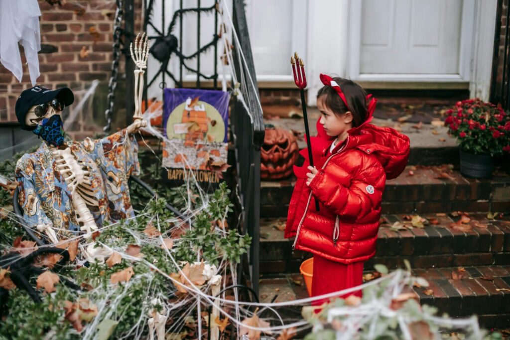 A child in a devil costume interacts with Halloween decorations, including a skeleton, on a decorated doorstep.