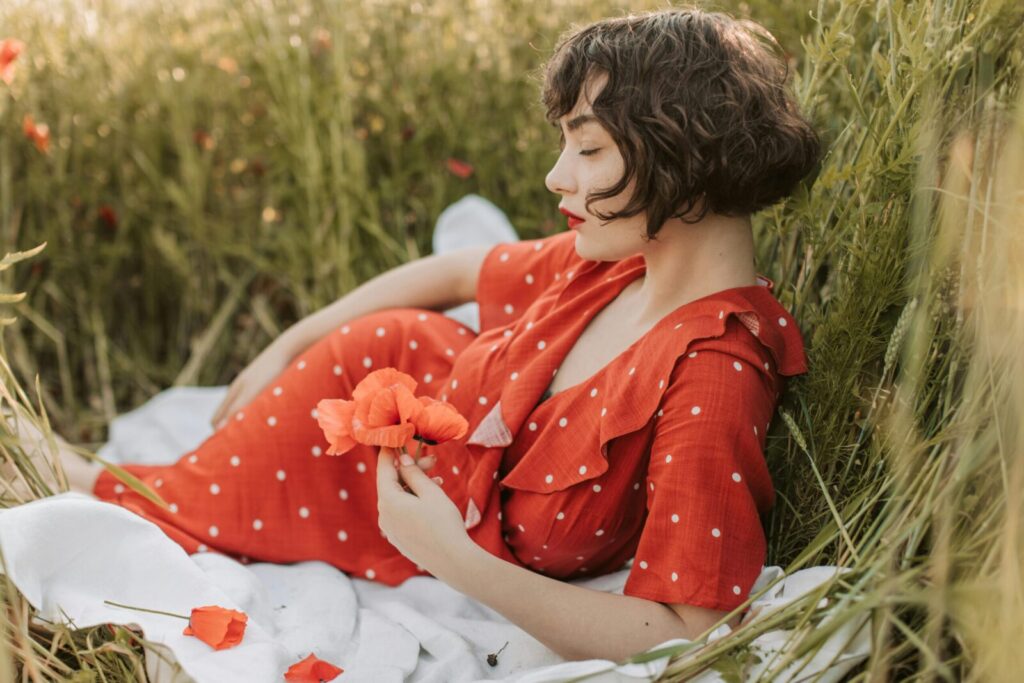 A woman in a red polka dot dress relaxes in a meadow holding poppies, creating a peaceful scene.