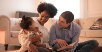 Loving parents reading with cheerful toddler in cozy living room, embracing family time.
