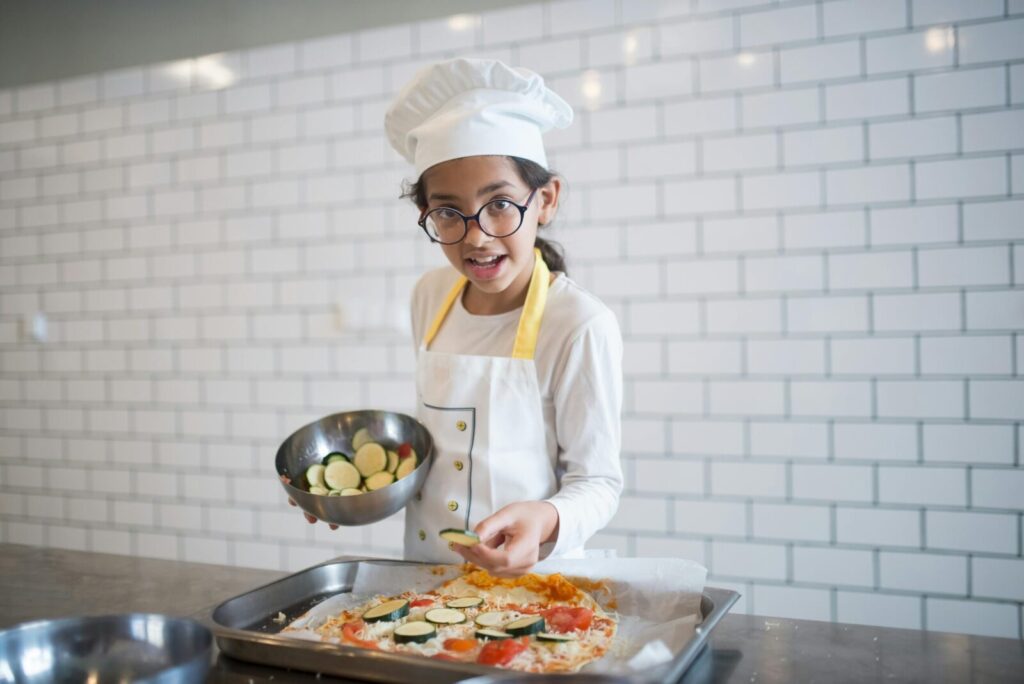 A young girl wearing a chef hat prepares pizza with fresh vegetables in a modern kitchen setting.