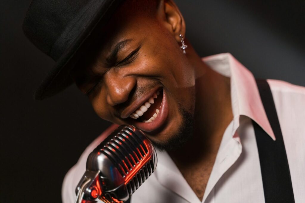 African American man passionately singing into a vintage microphone wearing a hat and white shirt.