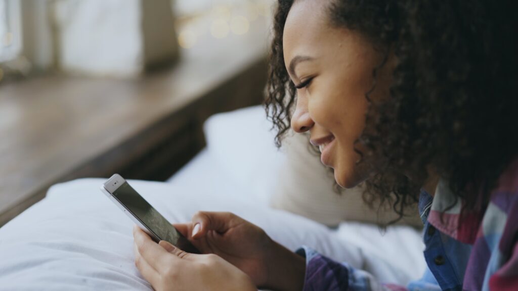 Young woman smiling while using her smartphone in bed.