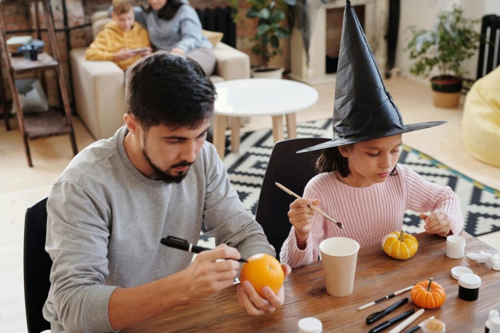 A father and daughter painting pumpkins together indoors, celebrating Halloween creativity.