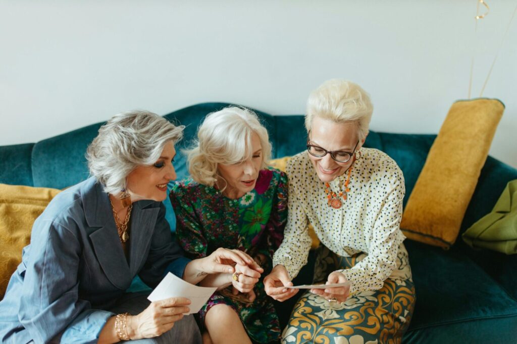 Three senior women joyfully reminiscing over old photos on a cozy couch.