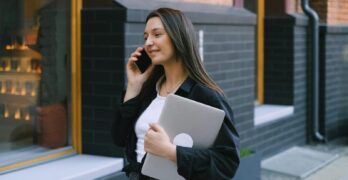 Confident woman walking down the street, making a phone call while holding a laptop.