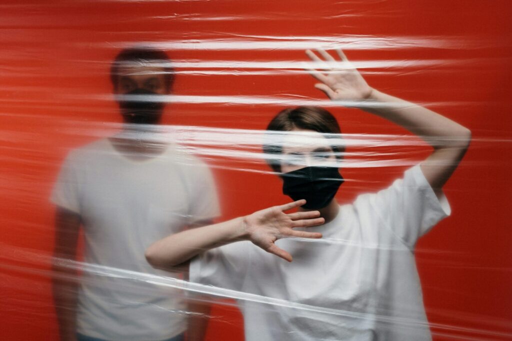 Two adults wearing masks stand behind a plastic sheet, symbolizing isolation and protection.