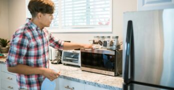 A man is cleaning a microwave in his modern kitchen using a spray bottle and cloth.