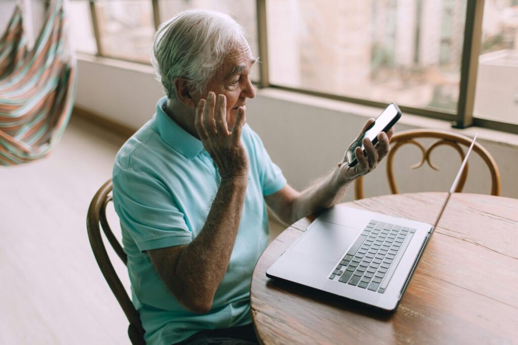 Elderly man using smartphone and laptop indoors, looking puzzled, showcasing technology and leisure.