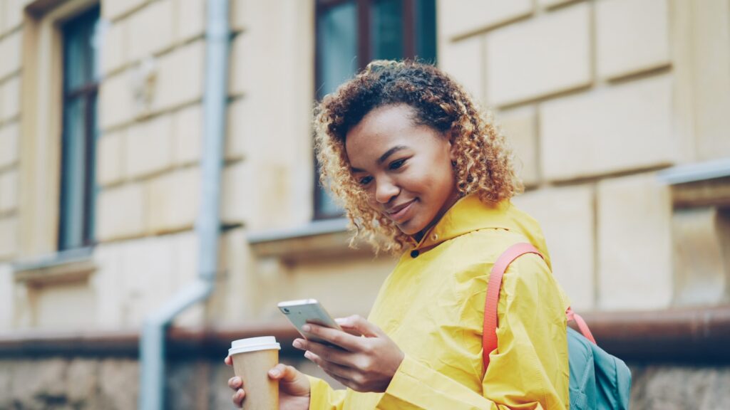 Young woman with coffee and phone walks outside.