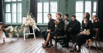 Mourners sitting indoors at a funeral ceremony, dressed in black, holding flowers.