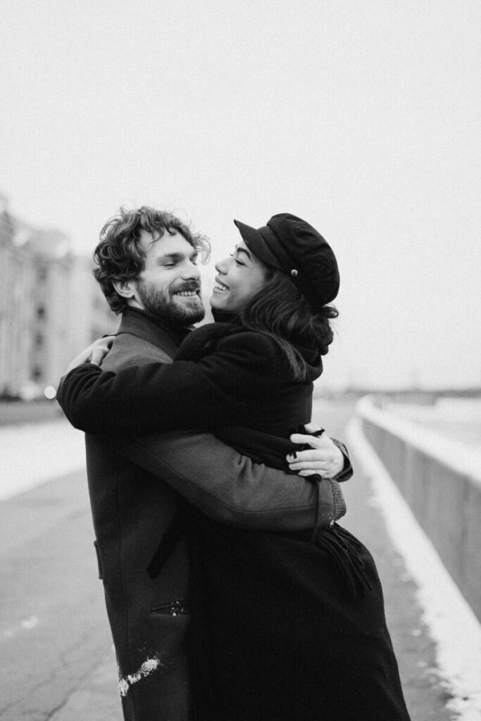 A joyful couple embraces on a snowy day, capturing love and happiness in black and white.