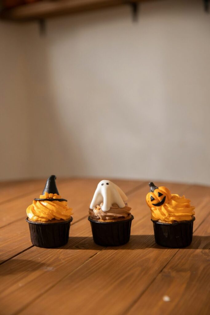 Three Halloween cupcakes featuring witch hat, ghost, and pumpkin designs on a wooden table.