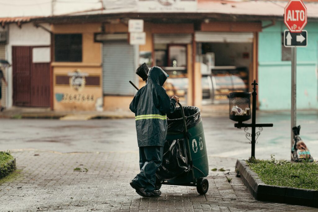 Sanitation worker pushing trash cart on wet street.