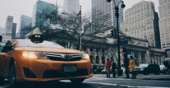 A bustling New York City street intersection featuring a yellow taxi and pedestrians with skyscrapers in the background.