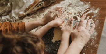 A parent and child bonding while kneading dough in a messy kitchen environment.