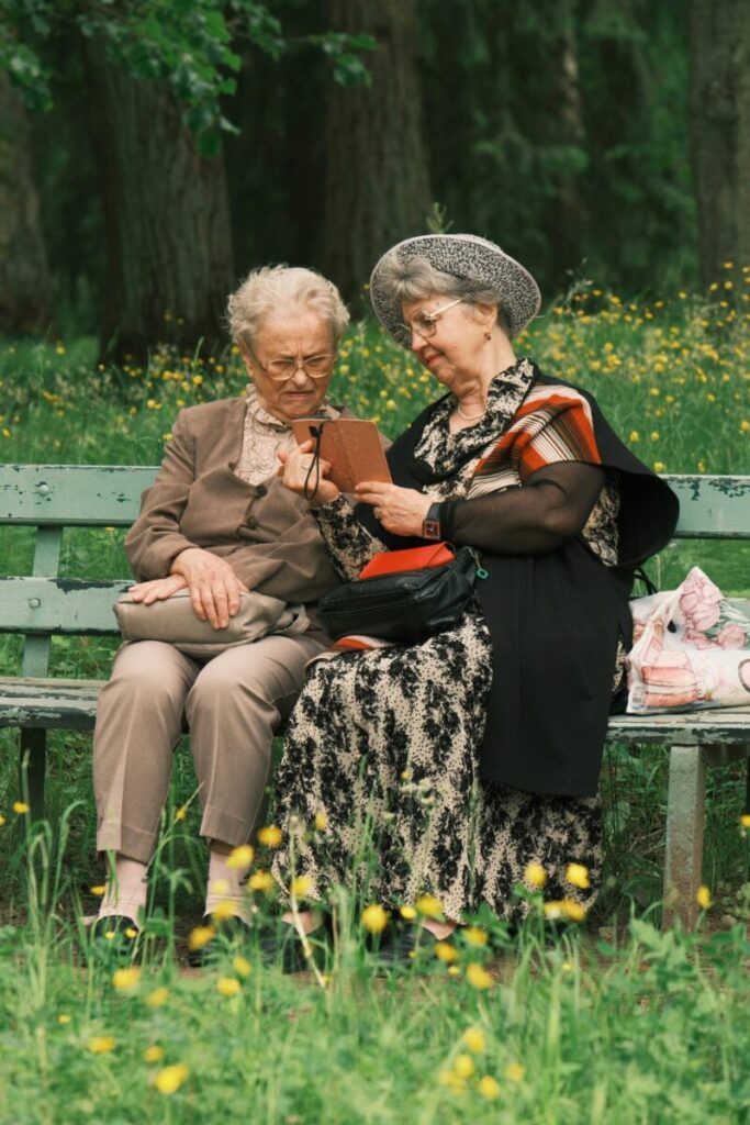 Two senior women engrossed in a book on a sunny park bench.