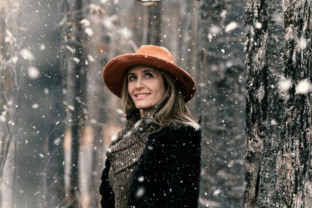 A joyful woman in a winter forest, wearing a stylish hat and coat during snowfall.