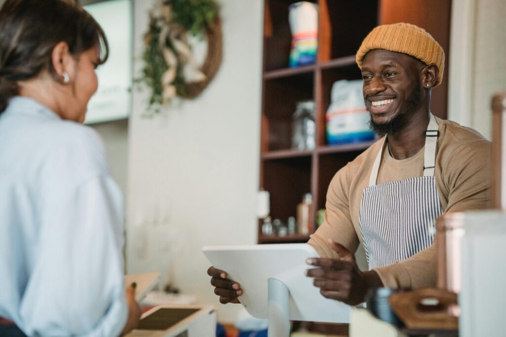 Barista with apron and beanie smiling as he takes an order from a customer in a cozy café setting.