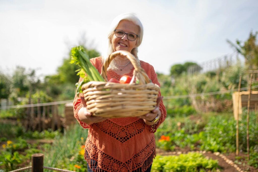 Elderly woman smiling with basket of fresh vegetables in a vibrant outdoor garden.