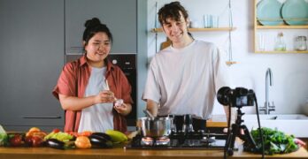 Two vloggers cooking and filming culinary content in a stylish kitchen setup.