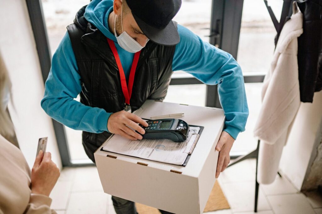 Delivery worker with face mask handling a package and credit card payment in a modern indoor setting.