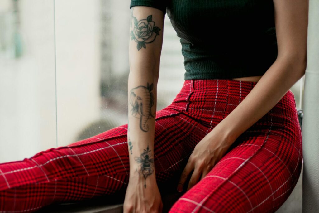 Stylish woman with tattoos poses by a window in red plaid pants, showcasing modern fashion.