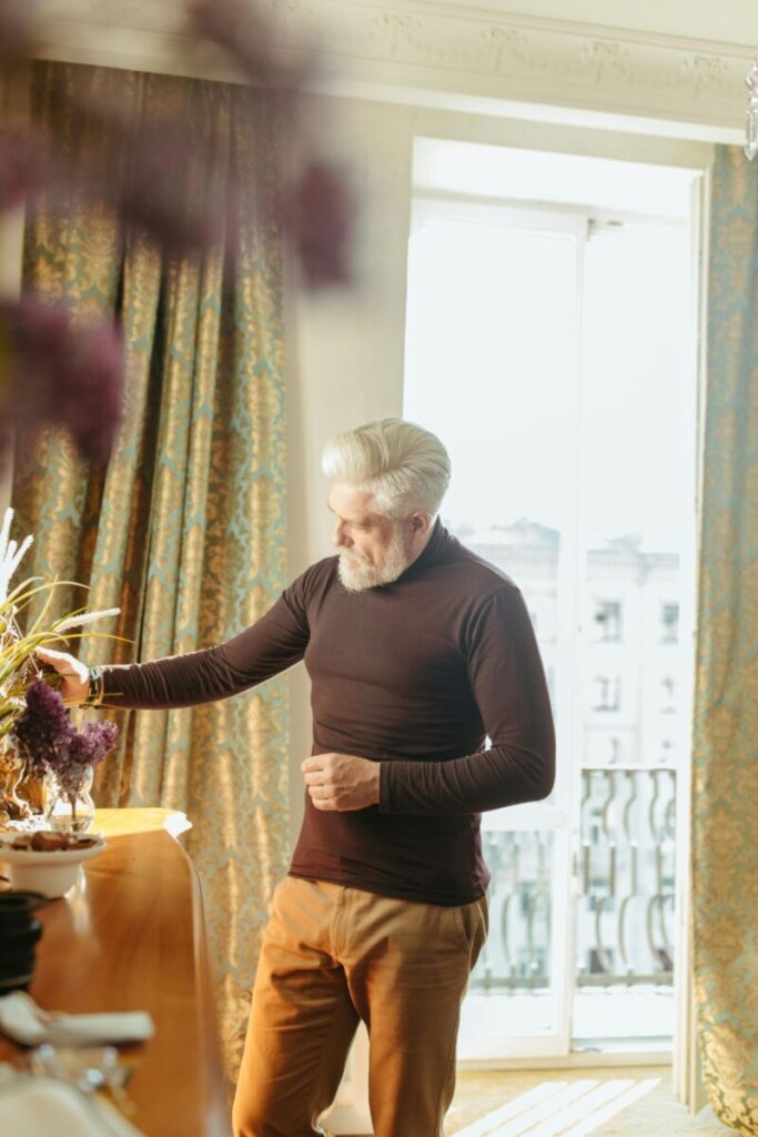 A senior man with a beard standing indoors, enjoying a bright morning light by the window.