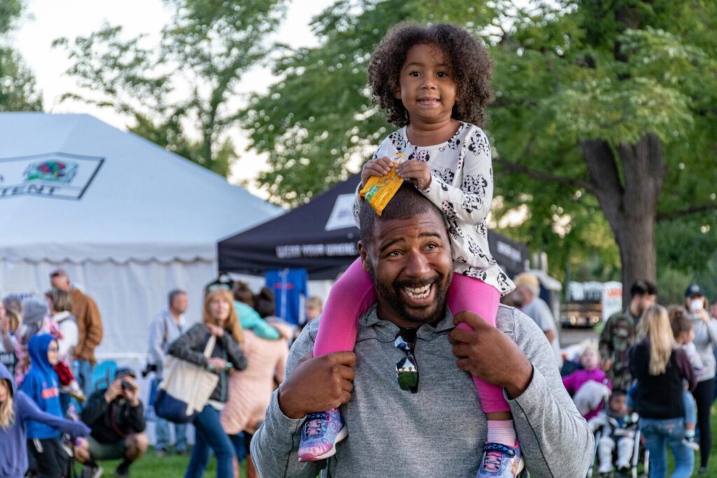 A joyful father carrying his daughter at an outdoor festival surrounded by people and tents.