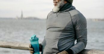 Mature man in activewear stands by the water, holding a tumbler and smiling.