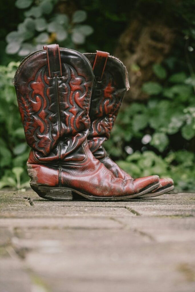 A pair of well-worn vintage cowboy boots resting on a rustic stone path with greenery in the background.