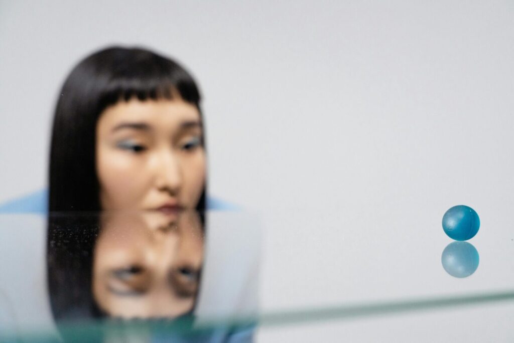 A woman gazes thoughtfully at a blue ball with reflections on a glass surface.