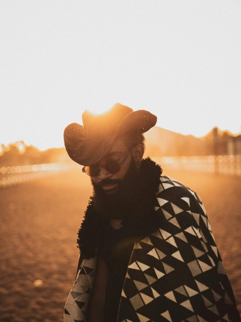 A fashionable man in a cowboy hat and patterned poncho at sunset in a Nigerian landscape.