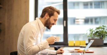 Man in office with coffee, smiling while working at laptop, captures the essence of remote work.