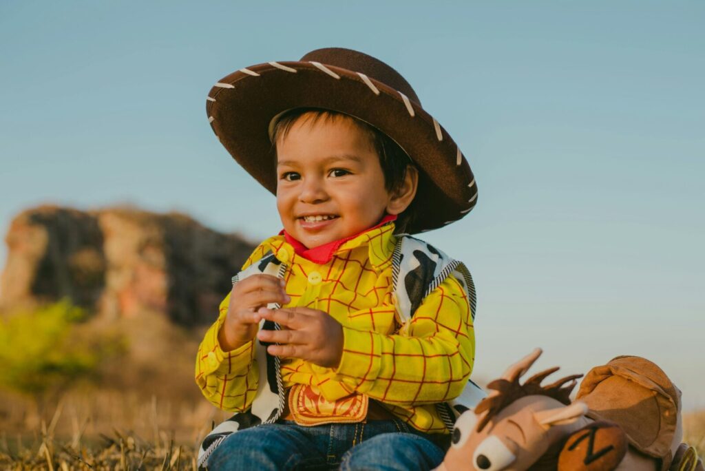 Smiling child dressed as a cowboy, holding stuffed animal toy in sunny outdoor setting