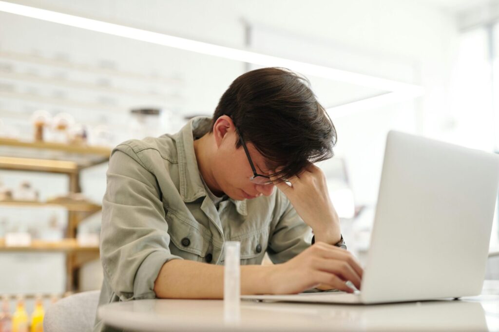 Exhausted young man working on a laptop inside a bright room, displaying fatigue.