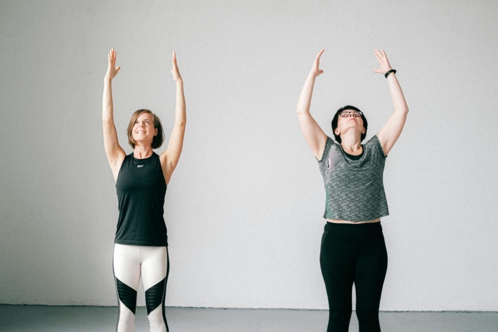 Two women engaged in a yoga session indoors, standing with arms raised, promoting fitness and wellness.
