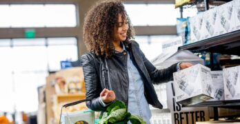 A woman carrying a grocery basket of vegetables picks up a Boxed Water box