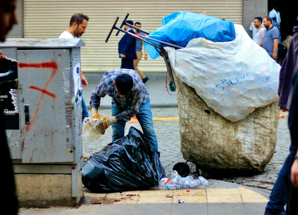A garbage collector cleaning up on a busy street with large trash bags and a cart.