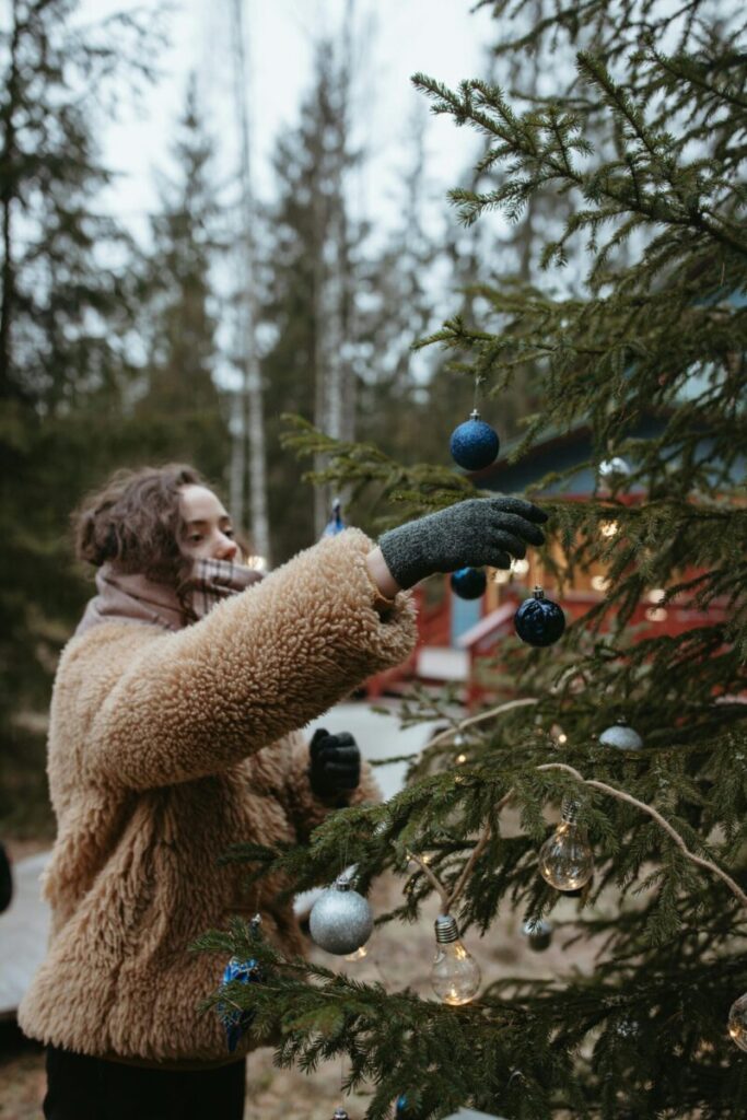 A woman in a cozy coat decorates a Christmas tree outdoors in a winter setting.