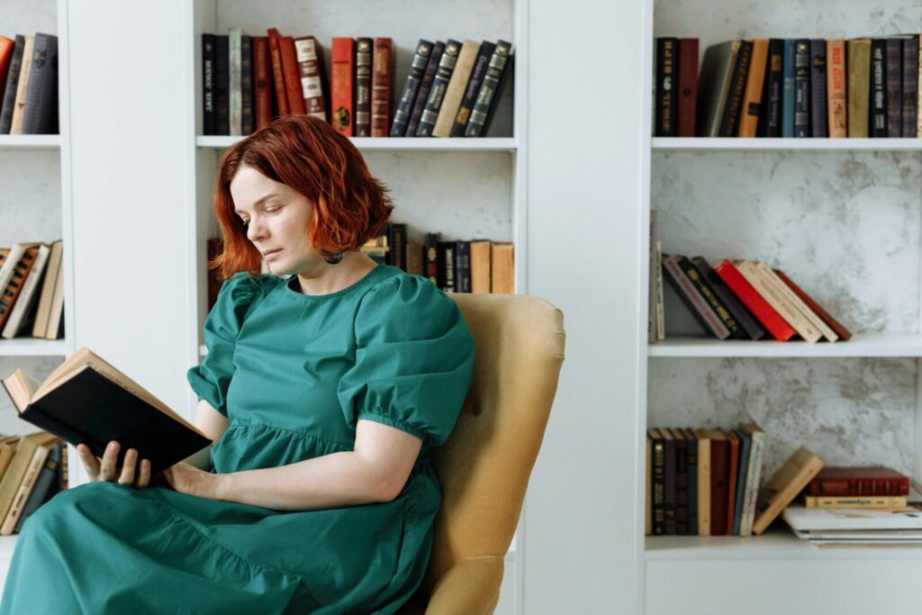 Woman reading a book while sitting in a library chair surrounded by bookshelves.