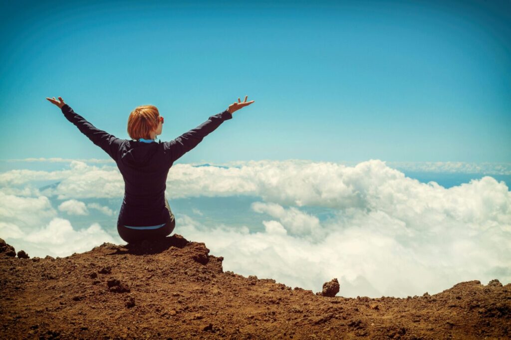 A woman enjoys a scenic view atop a cliff in Kula, Hawaii, surrounded by clouds and blue sky.
