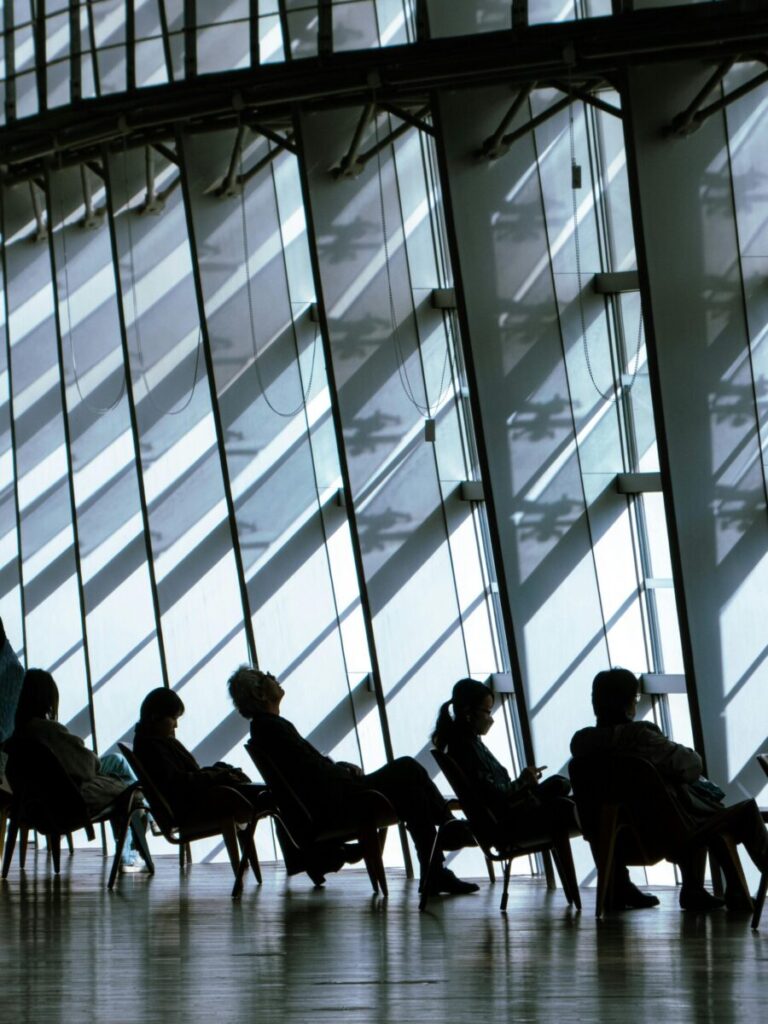Group of people sitting in chairs against large modern glass windows creating striking shadows.