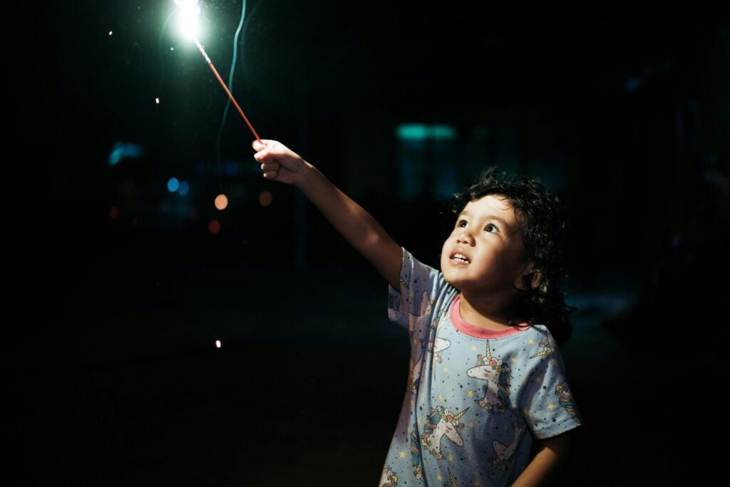 A young girl delightfully holds a sparkler, lighting up her face under the night sky.