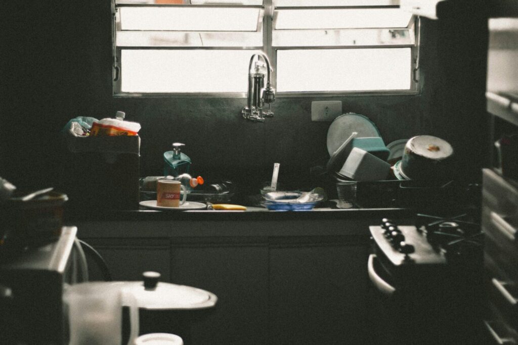 A cluttered kitchen sink filled with dirty dishes and utensils near a window, creating a messy home interior scene.